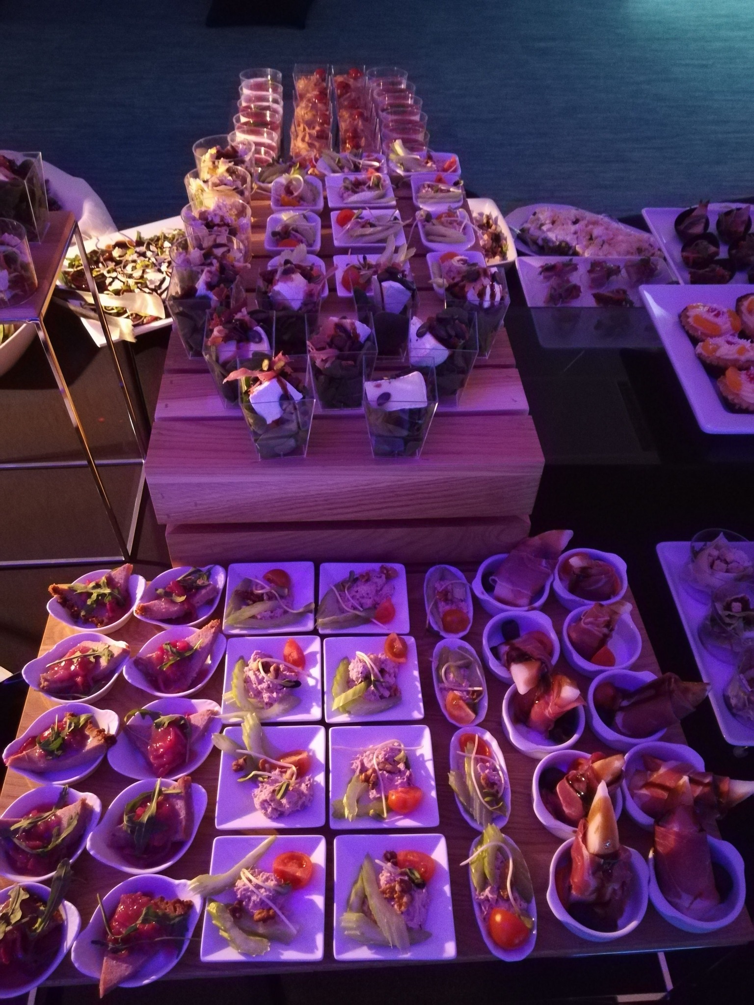 Bogato zastawiony stół z różnorodnymi przekąskami cateringowymi, w tym sałatki w szklanych naczyniach, tartinki i przystawki z szynką parmeńską, podświetlone fioletowym światłem.
