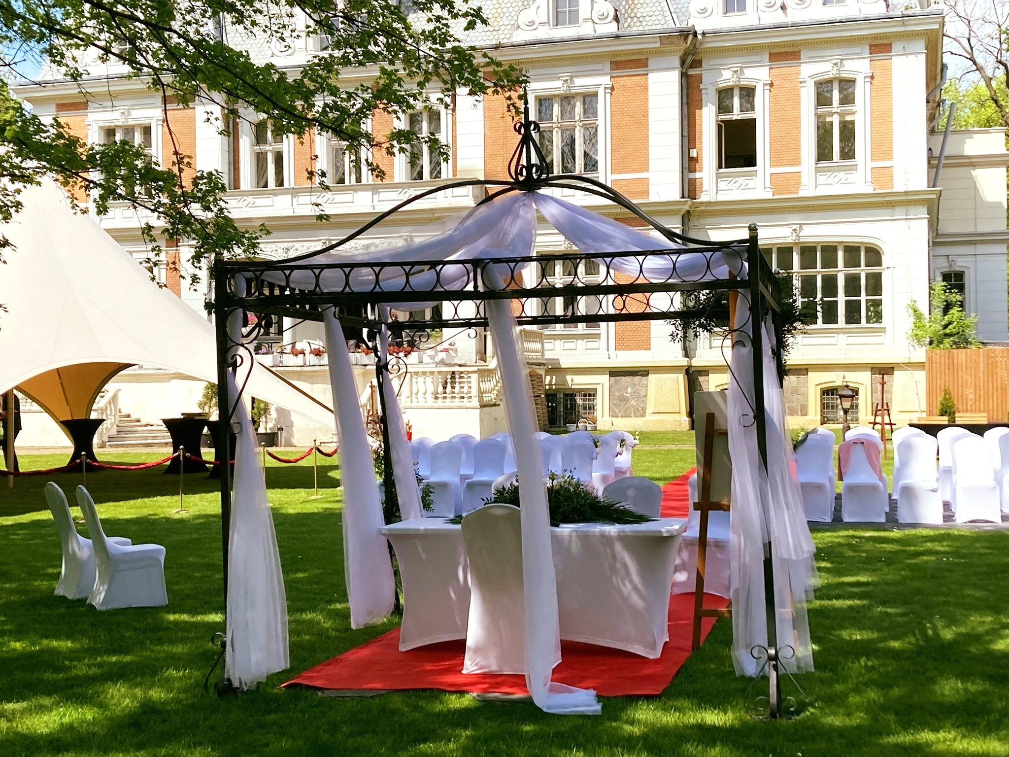 Uroczysta aranżacja ślubna w plenerze: metalowa altana z białymi draperiami, czerwony dywan, białe krzesła z dekoracjami, na trawniku przed budynkiem.