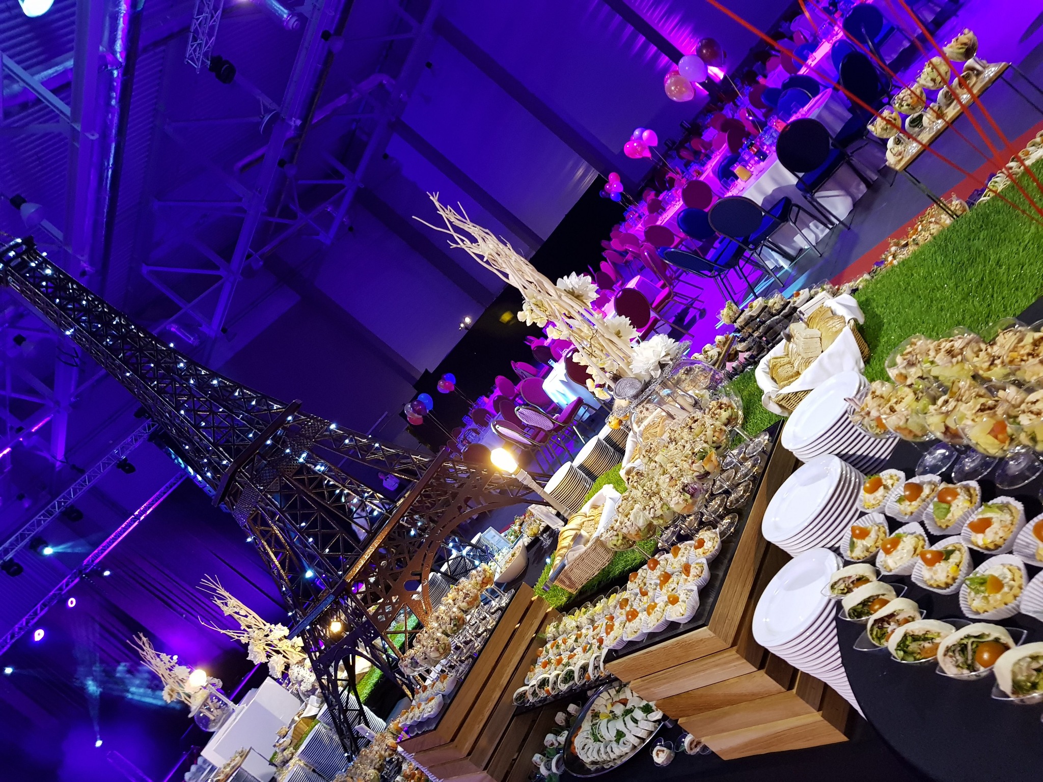 Ukośne ujęcie stołu cateringowego z przekąskami, dekoracjami kwiatowymi i miniaturą wieży Eiffla pod fioletowym oświetleniem sali eventowej.