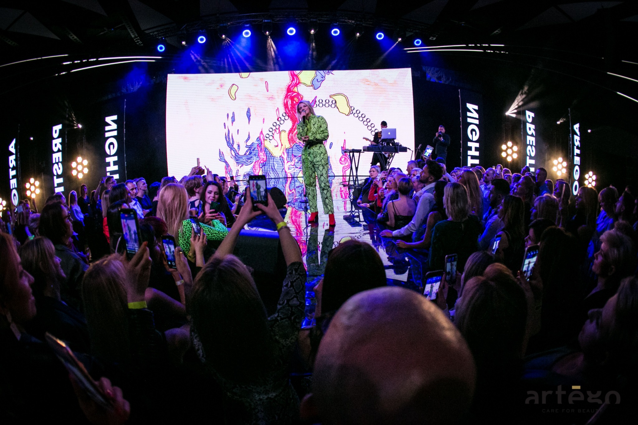 Wokalistka w zielonym kombinezonie wężowym i czerwonych butach na scenie podczas koncertu, oświetlona reflektorami, tłum publiczności filmuje występ telefonami, w tle duży ekran LED z abstrakcyjną...