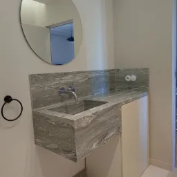 Łazienka z blatem kamiennym o marmurkowym wzorze, okrągłe lustro, czarny wieszak na ręczniki. Nowoczesne wykończenie, jasne kolory, minimalistyczny design.