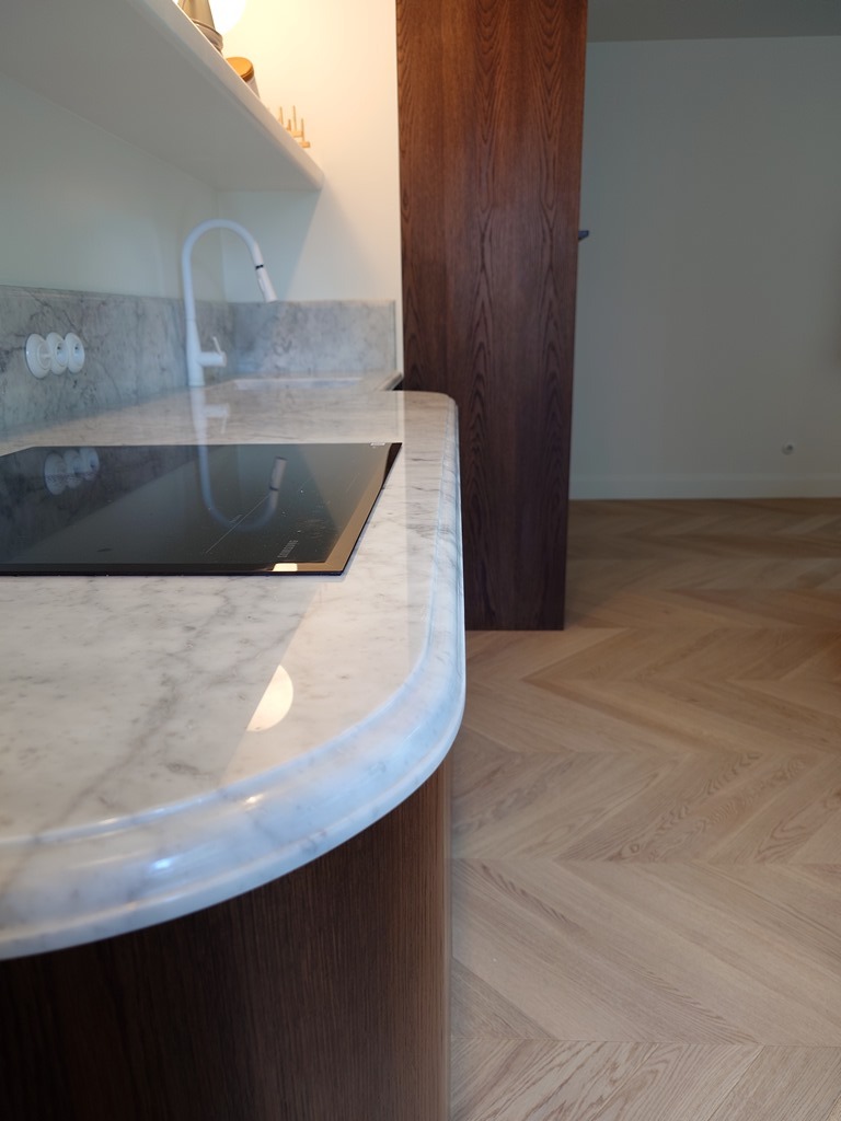 Marmurowy blat kuchenny z zaokrąglonym brzegiem i wbudowaną płytą grzewczą, na tle drewnianej zabudowy i parkietu ułożonego w jodełkę.