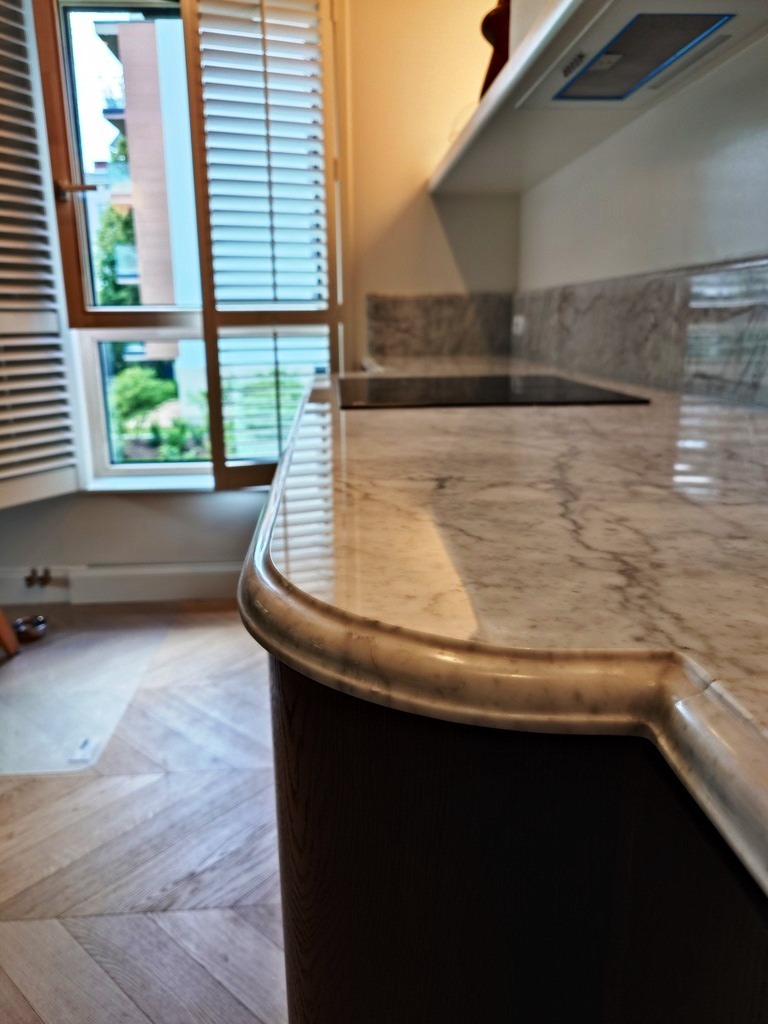 Elegancki blat kamienny z marmurowym wzorem w jasnej kuchni z drewnianą podłogą i oknem z żaluzjami w tle. Zaokrąglony brzeg blatu.