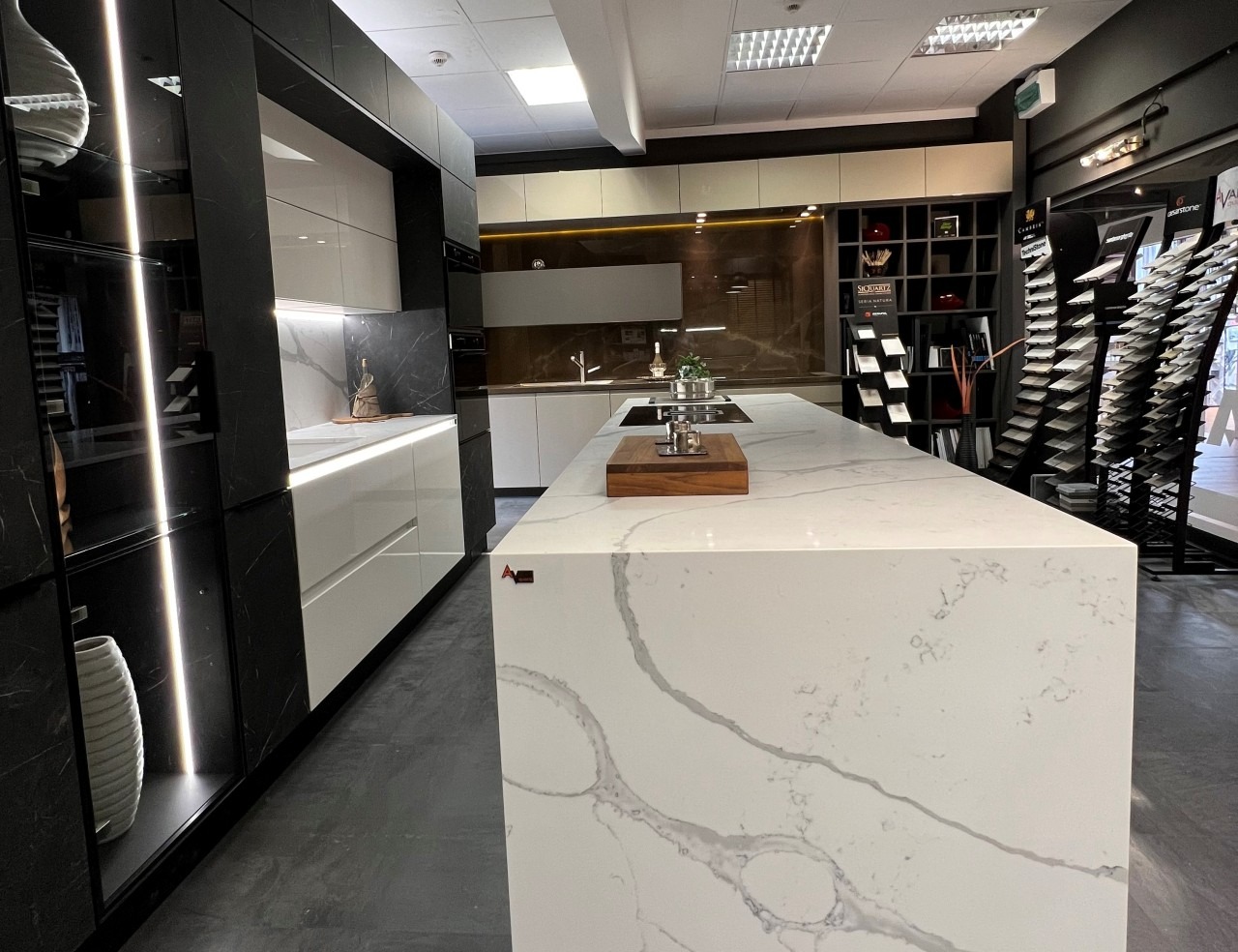 Nowoczesna kuchnia z wyspą i blatem z białego kamienia z szarymi żyłami. W tle ekspozycja próbek blatów. Minimalistyczny design z oświetleniem LED.