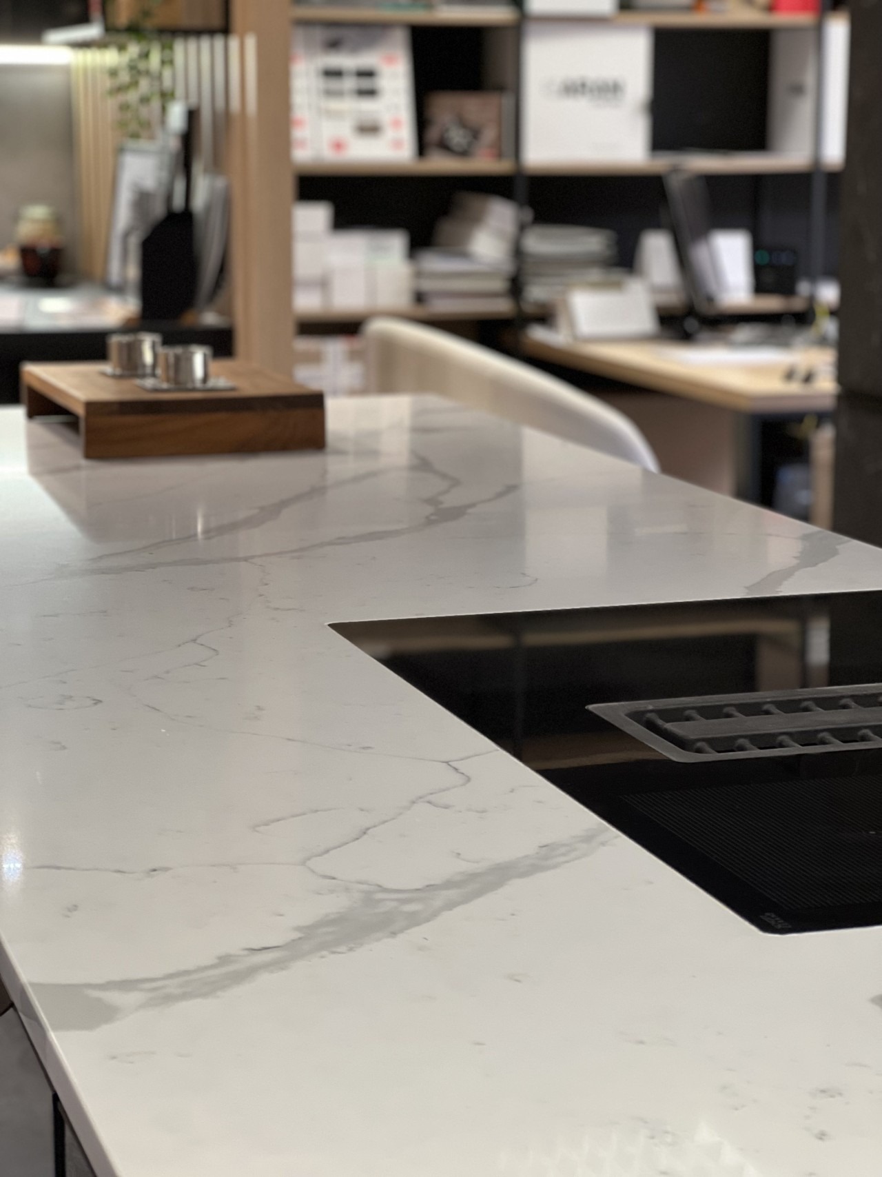 Biały blat kuchenny z marmurowym wzorem i wbudowaną czarną płytą grzewczą. Na blacie drewniana podstawka z metalowymi elementami dekoracyjnymi.