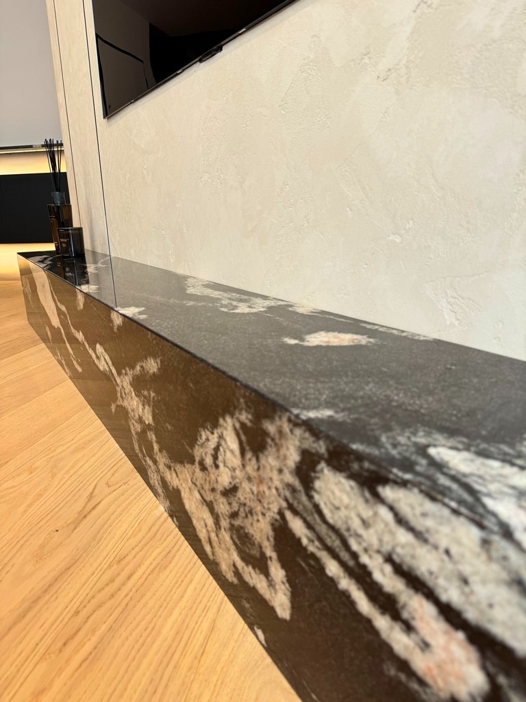 Długi, czarny blat z wzorem, umieszczony pod telewizorem na ścianie o fakturze. Podłoga drewniana. Blat z kamienia naturalnego.