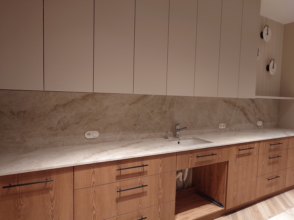 Jasny blat z marmurowym wzorem w nowoczesnej kuchni z drewnianymi szafkami i minimalistycznymi uchwytami. Widoczne gniazdka elektryczne i dekoracyjne lampy ścienne.