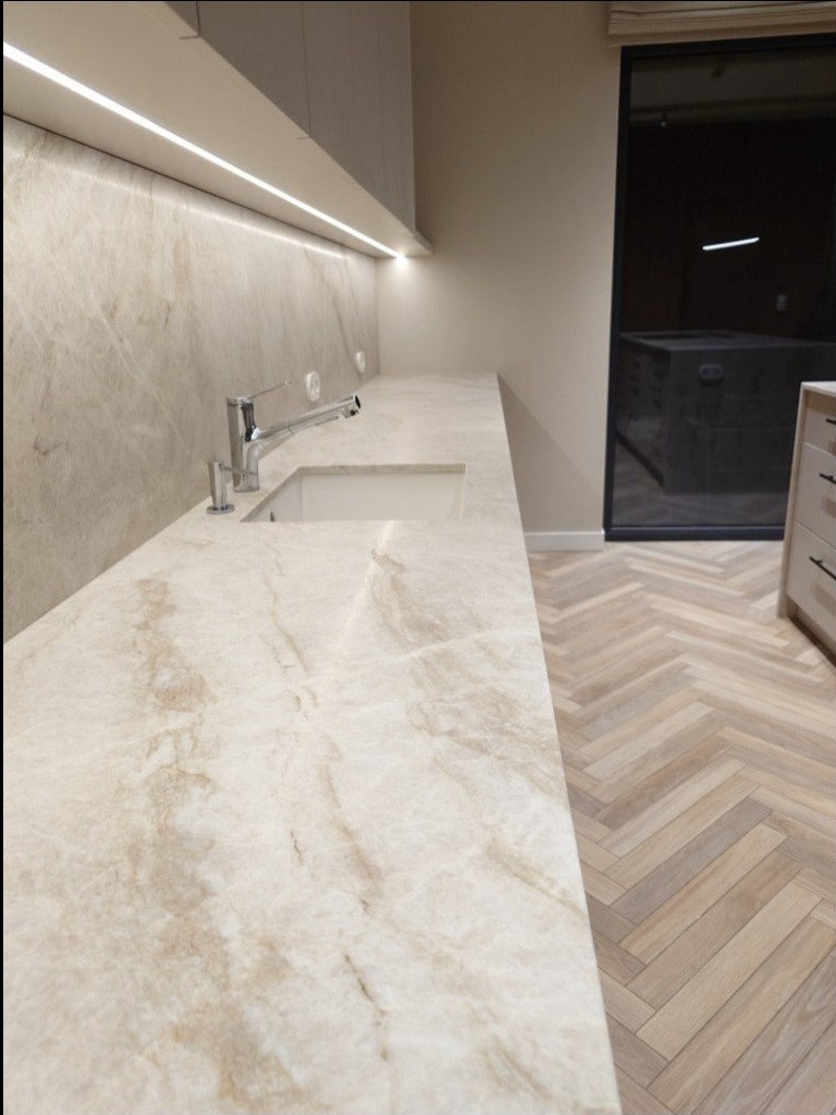 Beżowy blat kamienny z umywalką i baterią w nowoczesnej kuchni z oświetleniem LED pod szafkami i parkietem w jodełkę. Widok z perspektywy.