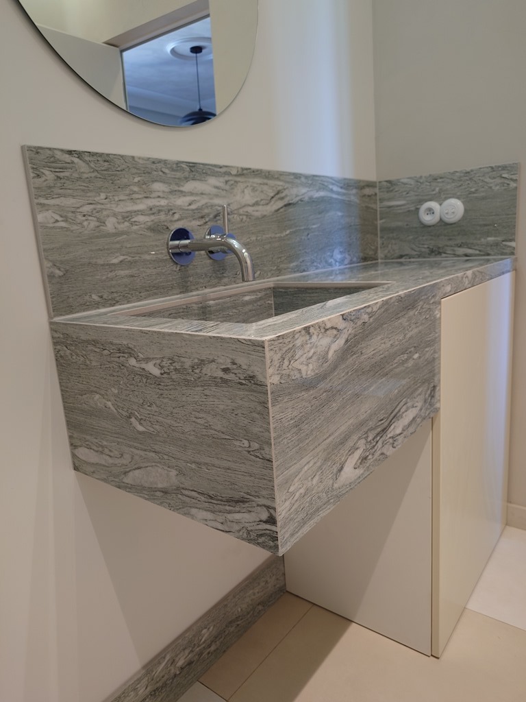 Szara umywalka z kamienia o wyraźnym wzorze żyłkowania, w nowoczesnej łazience z białymi szafkami i okrągłym lustrem. Widoczne gniazdko elektryczne na ścianie.