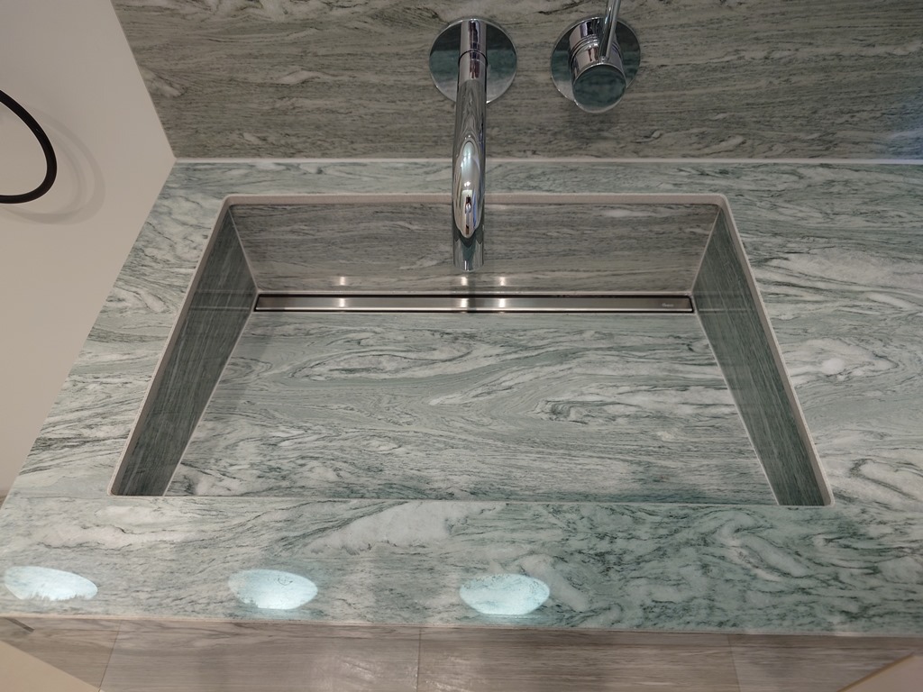 Szaro-zielony blat z kamienia naturalnego z wbudowaną umywalką i chromowaną armaturą. Widoczny liniowy odpływ w umywalce i subtelne oświetlenie punktowe w blacie.