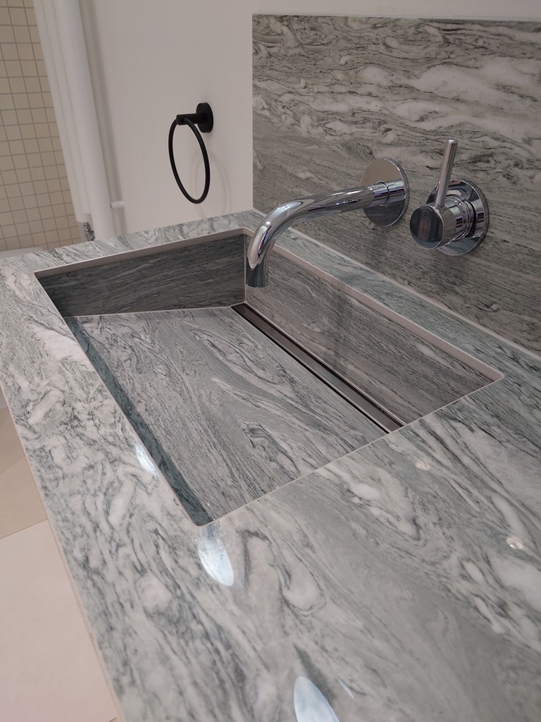 Szary blat z kamienia z wbudowaną umywalką i chromowaną armaturą, widoczny w łazience. W tle biała ściana z czarnym uchwytem na ręcznik.