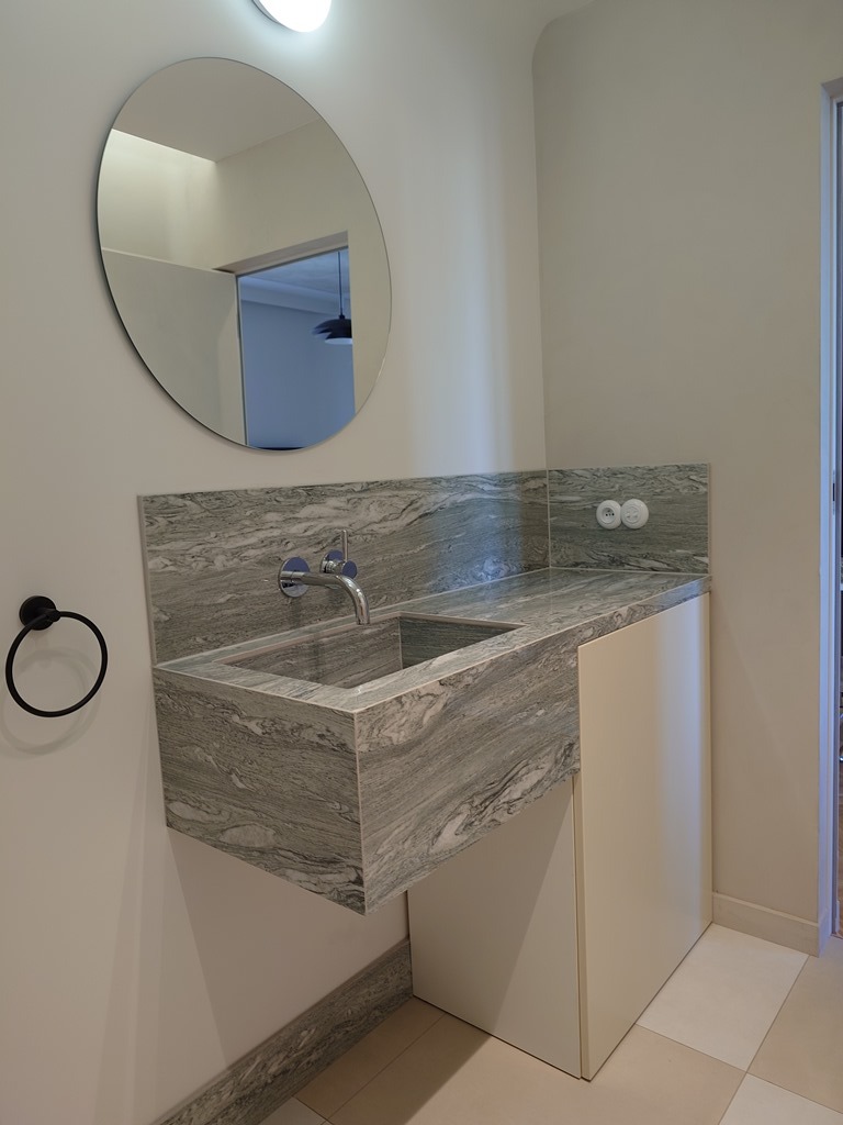 Łazienka z blatem kamiennym o marmurkowym wzorze, okrągłe lustro, czarny wieszak na ręczniki. Nowoczesne wykończenie, jasne kolory, minimalistyczny design.