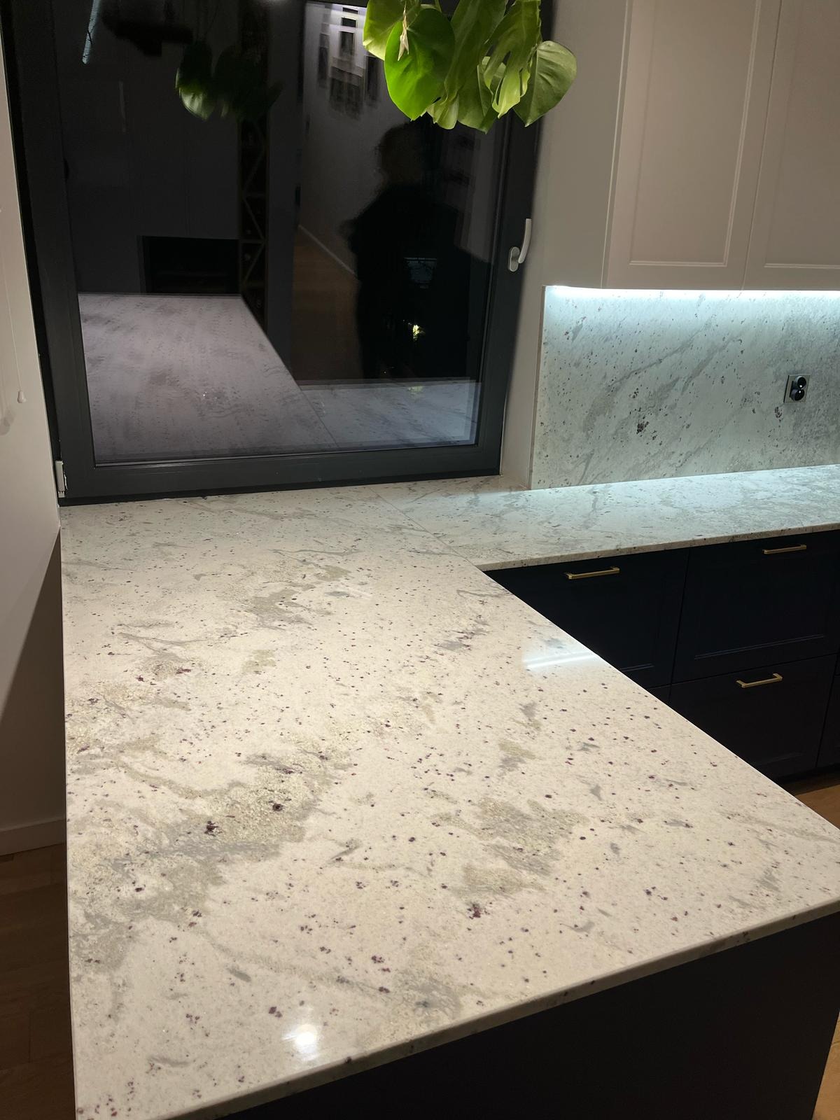 Jasny blat z granitu Kashmir White w kuchni, z widocznym oknem i rośliną doniczkową. Minimalistyczne, ciemne szafki z złotymi uchwytami. Podświetlenie LED na ścianie.