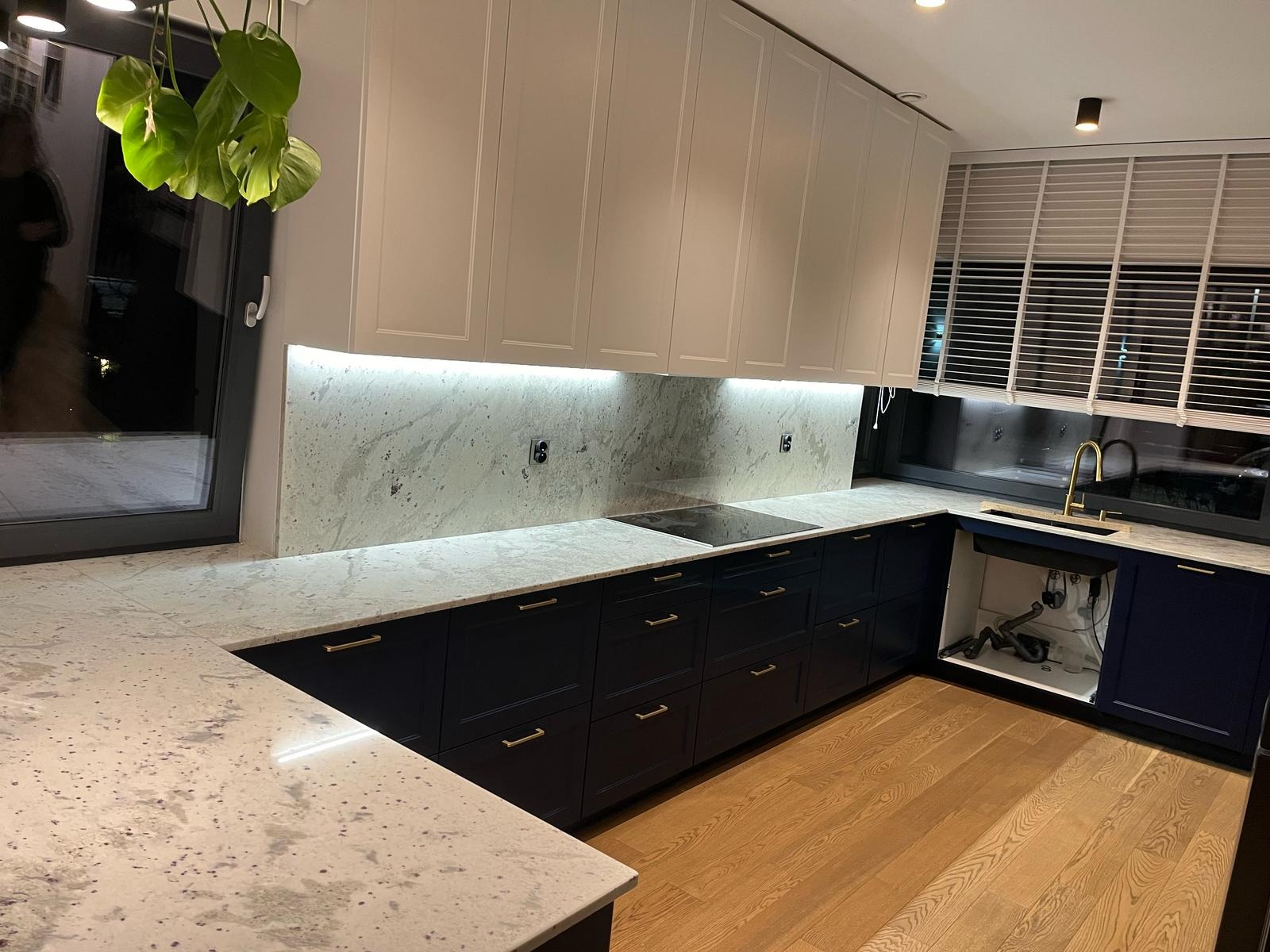 Jasny blat z kamienia w kuchni z ciemnymi szafkami i białymi górnymi szafkami. Nad blatem podświetlana ściana z tego samego materiału. Nowoczesny design.