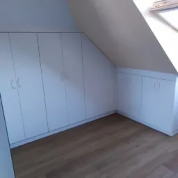 Białe szafy wnękowe z uchwytami, dopasowane do skosu poddasza, z widocznym oknem dachowym i drewnianą podłogą.