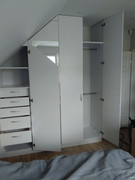 Biała szafa wnękowa z otwartymi drzwiami i szufladami, dopasowana do skosu dachu, z drewnianą podłogą i widocznym fragmentem łóżka.