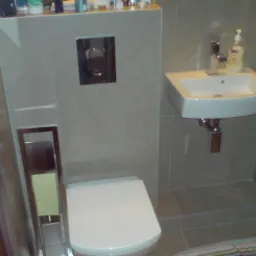 Nowoczesna łazienka z szarymi płytkami, podwieszaną toaletą z chromowanym przyciskiem spłukiwania, białą umywalką i chromowaną baterią, oraz częściowo widoczną kabiną prysznicową z pomarańczowym...