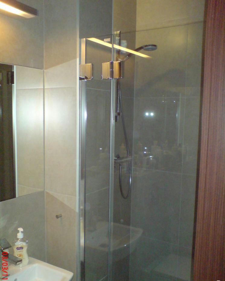 Szklana kabina prysznicowa z chromowanymi okuciami i deszczownicą, widoczna umywalka z dozownikiem mydła, szare płytki na ścianach, oświetlenie sufitowe.