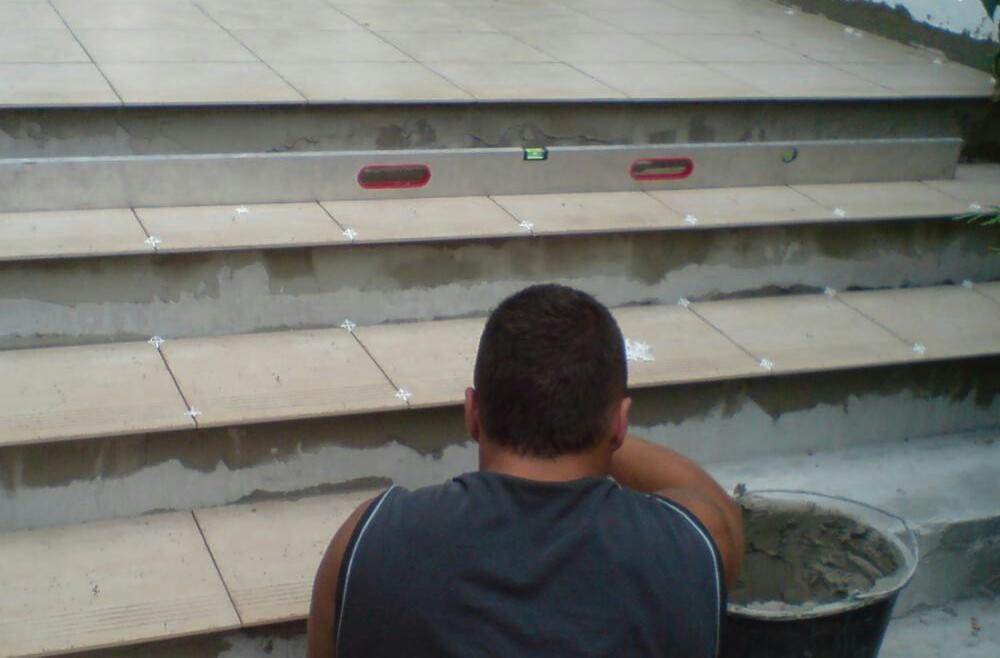 Mężczyzna w szarej koszulce układa płytki na schodach zewnętrznych, używając poziomicy i krzyżyków dystansowych, z widocznym wiadrem z zaprawą.