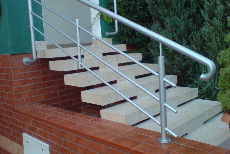 Zewnętrzne schody wyłożone jasnymi płytkami z zamontowaną, nowoczesną, srebrną balustradą z rurek.