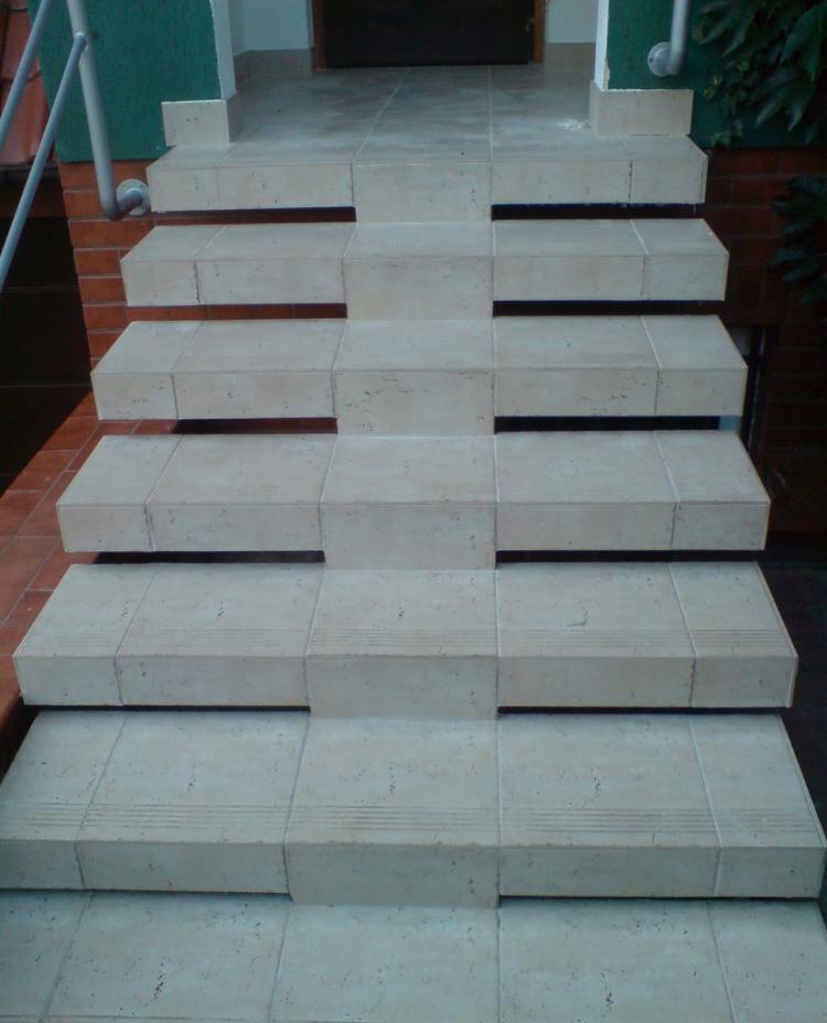 Schody zewnętrzne wyłożone jasnymi, kwadratowymi płytkami ceramicznymi z delikatnym wzorem, prowadzące do drzwi wejściowych, z widoczną metalową barierką po lewej stronie i fragmentem ceglanej...