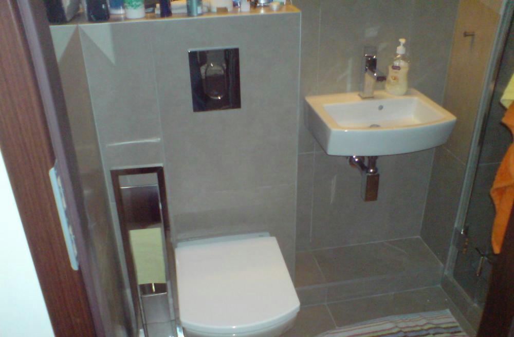 Nowoczesna łazienka z szarymi płytkami, podwieszaną toaletą z chromowanym przyciskiem spłukiwania, białą umywalką i chromowaną baterią, oraz częściowo widoczną kabiną prysznicową z pomarańczowym...