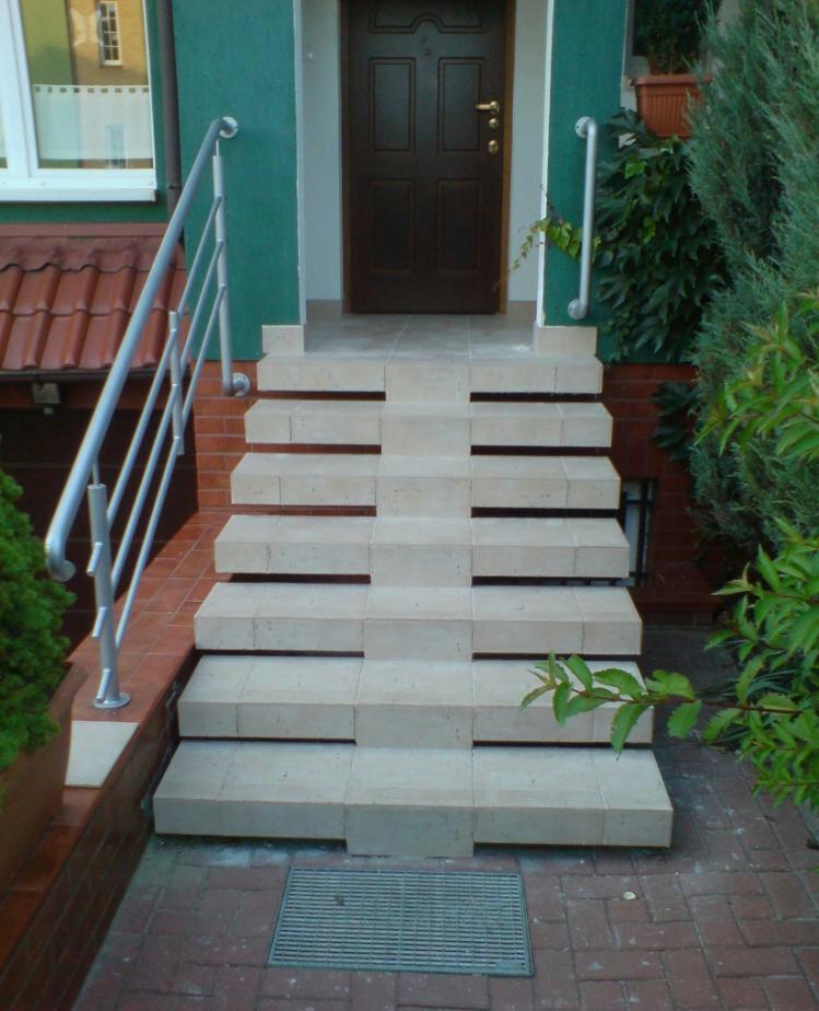 Schody wejściowe wyłożone jasnymi płytkami z metalową kratką odpływową u podstawy, prowadzące do ciemnobrązowych drzwi wejściowych w zielonej fasadzie budynku.