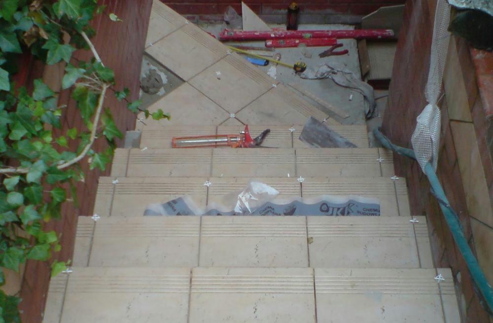 Kładzenie płytek na schodach zewnętrznych w Bytomiu, widoczne narzędzia glazurnicze i krzyżyki dystansowe, częściowo zakończony montaż, z bliska widoczne logo producenta chemii budowlanej.