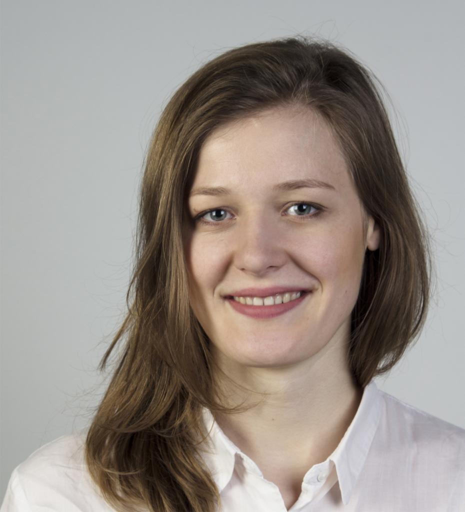 Portret uśmiechniętej kobiety z długimi brązowymi włosami i niebieskimi oczami w białej koszuli na jasnoszarym tle.