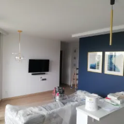 Pokój po malowaniu z granatową ścianą, zabezpieczone meble folią, widoczny odkurzacz przemysłowy i puszka z farbą Designer White na blacie