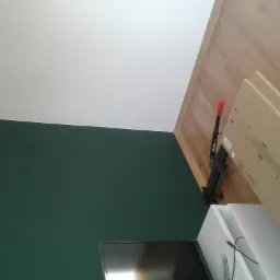 Świeżo pomalowana ściana w odcieniach butelkowej zieleni, kontrastująca z białym sufitem i jasną drewnianą podłogą, z widocznym fragmentem szafki RTV i narzędziem YATO z czerwoną rączką.