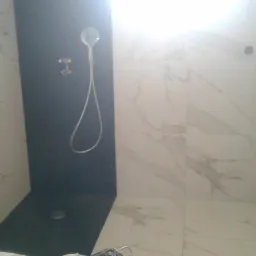 Ukończona kabina prysznicowa z czarną ścianą i deszczownicą, obłożona białymi płytkami imitującymi marmur, z widocznymi instalacjami wodnymi i odpływem liniowym.