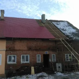 Częściowo wymieniony dach z czerwonej dachówki na starym ceglanym domu, widoczne drewniane łaty i rusztowania, śnieg na dachu i ziemi.