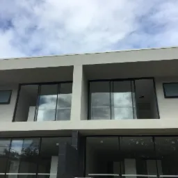 Dwukondygnacyjny budynek z dużymi przesuwnymi oknami, elewacja w jasnym odcieniu szarości, fragment nieba z chmurami w odbiciu szyb.
