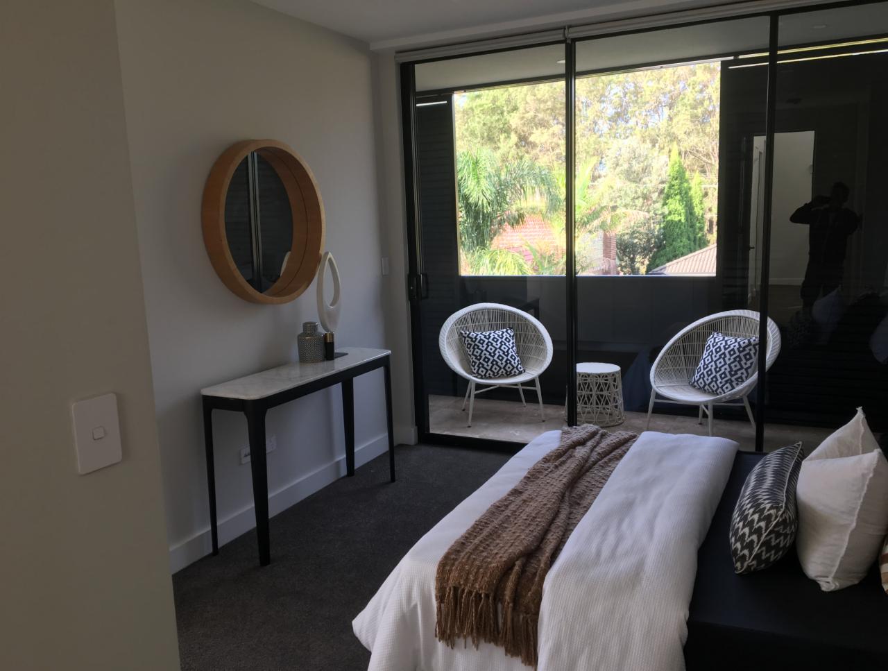 Jasne wnętrze sypialni z wyjściem na balkon, widoczne rattanowe krzesła z poduszkami w geometryczne wzory, konsola z okrągłym lustrem, oraz częściowo widoczne łóżko z narzutą w odcieniach beżu...