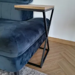 Stolik pomocniczy z drewnianym blatem i metalową podstawą w kształcie litery X, dostawiony do boku granatowej, welurowej sofy na tle parkietu i dywanu.