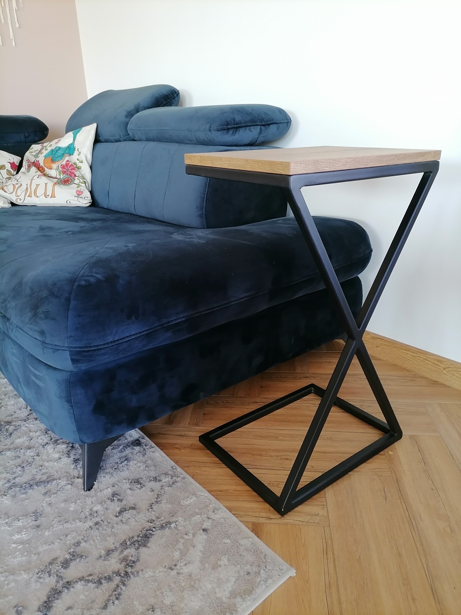 Stolik pomocniczy z drewnianym blatem i metalową podstawą w kształcie litery X, stojący obok granatowej, welurowej sofy z poduszką dekoracyjną