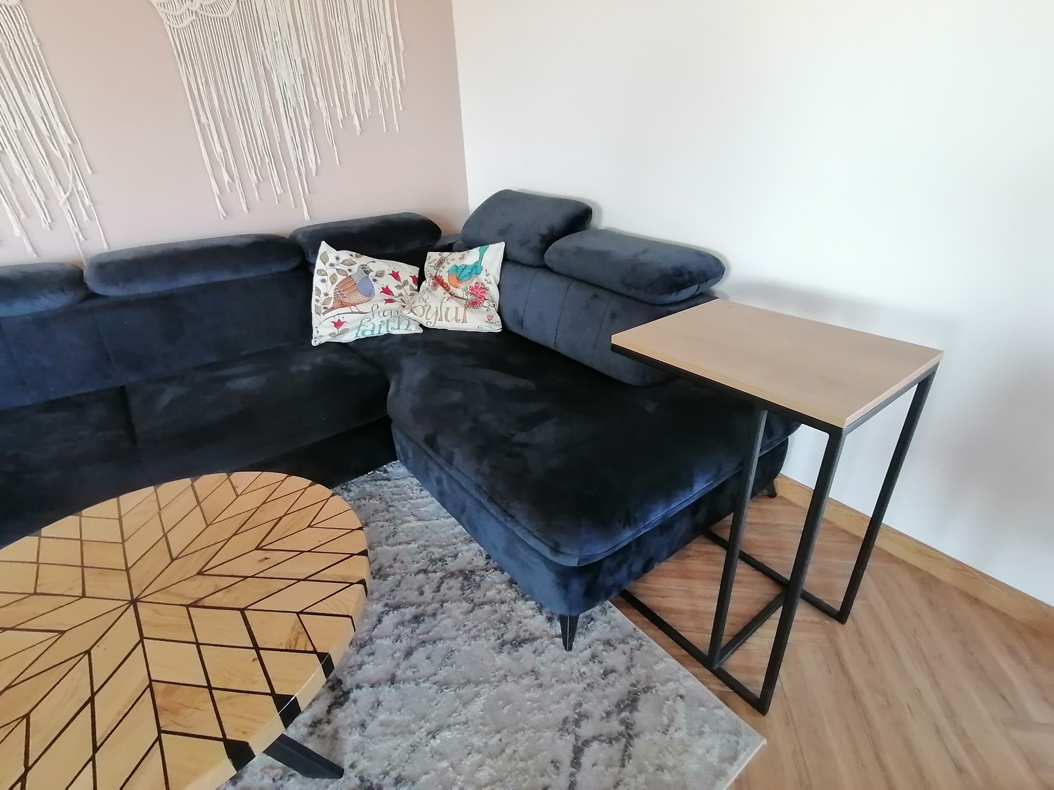 Wnętrze salonu z narożną, ciemnoniebieską sofą z poduszkami dekoracyjnymi, owalnym stolikiem kawowym z drewnianym blatem o geometrycznym wzorze i stolikiem pomocniczym z jasnym blatem i czarną...