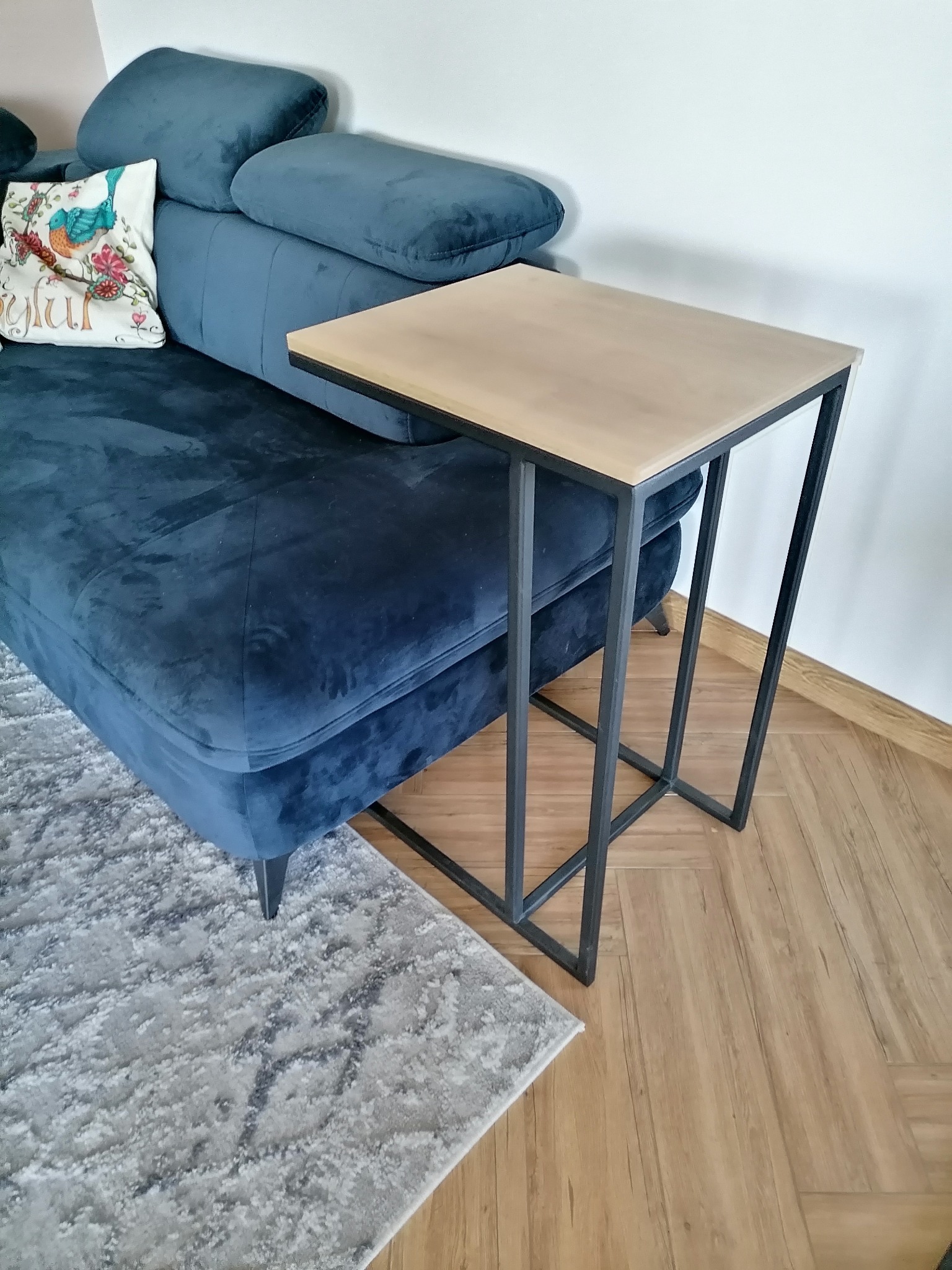 Stolik pomocniczy z blatem drewnianym i metalową, czarną ramą, ustawiony obok granatowej, welurowej sofy z dekoracyjną poduszką we wnętrzu z drewnianą podłogą i szarym dywanem.