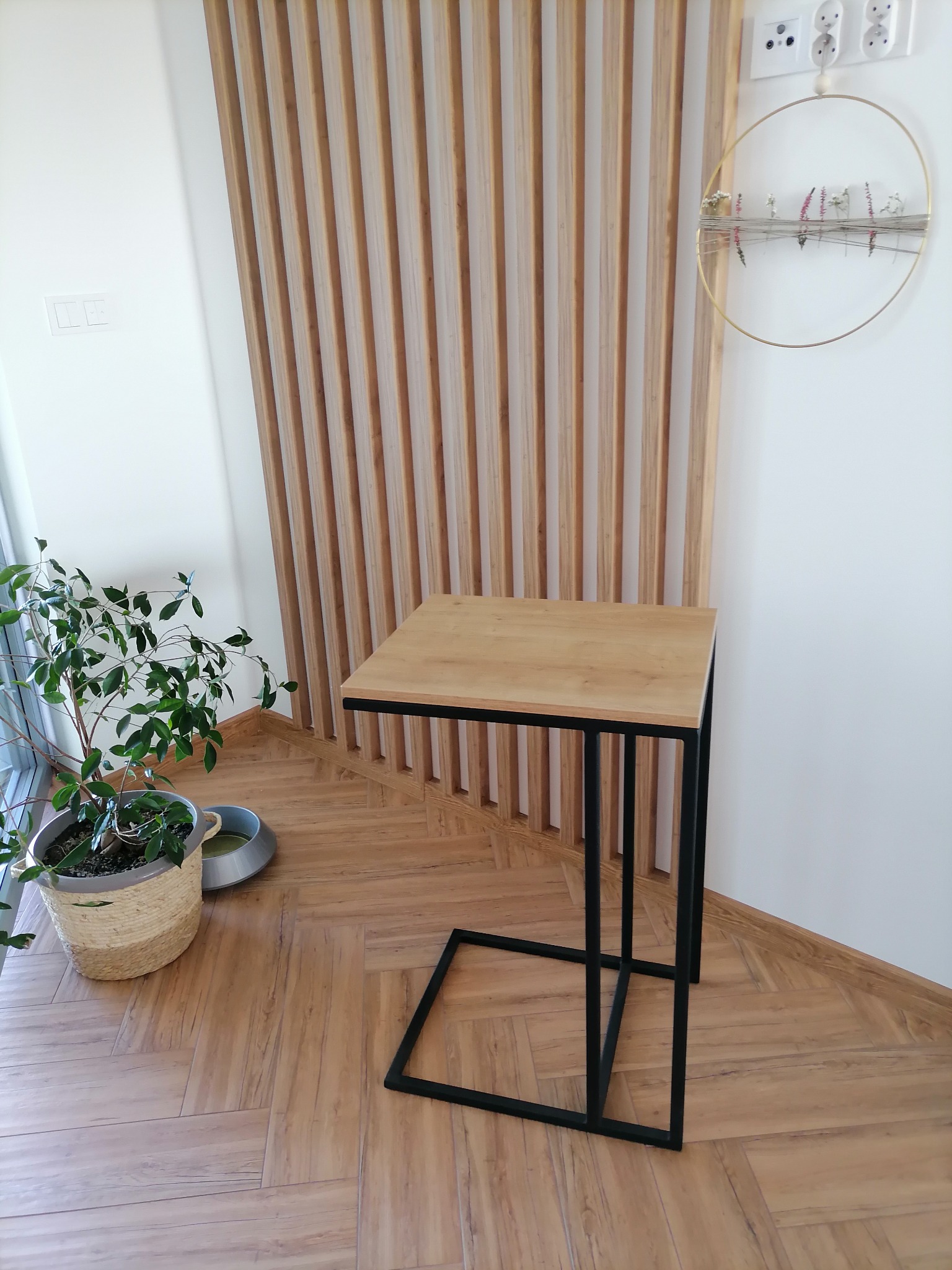 Stolik pomocniczy z blatem z jasnego drewna i czarną metalową podstawą w nowoczesnym wnętrzu z drewnianą ścianką działową i rośliną doniczkową.
