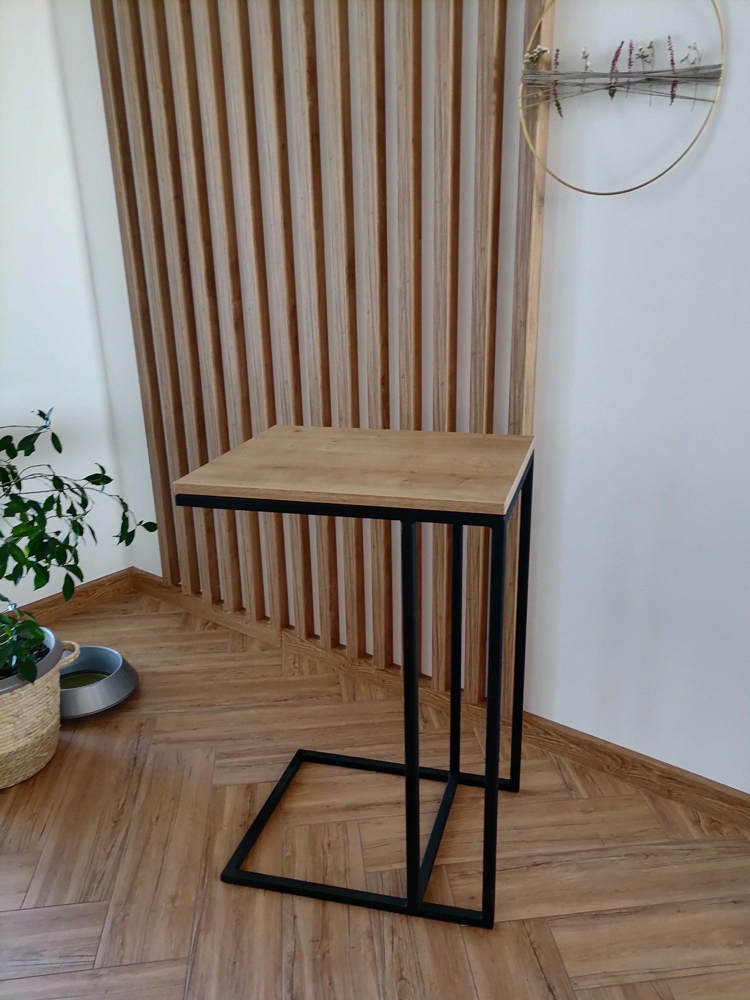 Stolik pomocniczy z drewnianym blatem i czarną, metalową ramą w minimalistycznym wnętrzu z drewnianą ścianką działową i okrągłą dekoracją ścienną z suszonych kwiatów.