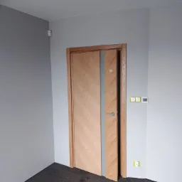 Drewniane drzwi wewnętrzne z przeszkleniem w szarej ścianie, widoczne gniazdka elektryczne i czujnik alarmowy.