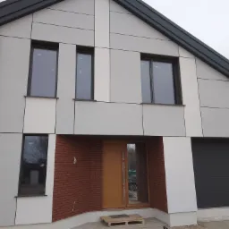 Nowoczesny dom jednorodzinny z elewacją z paneli w odcieniach szarości i bieli, akcentem z cegły klinkierowej przy wejściu i grafitową stolarką okienną, widok z zewnątrz w pochmurny dzień.