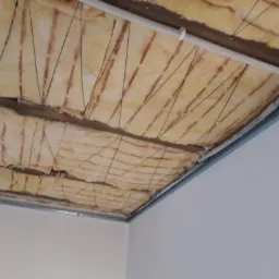 Izolacja termiczna poddasza z widocznymi belkami, wełną mineralną i metalowym profilem, przymocowana sznurkami do konstrukcji dachu, przed zakryciem płytami.