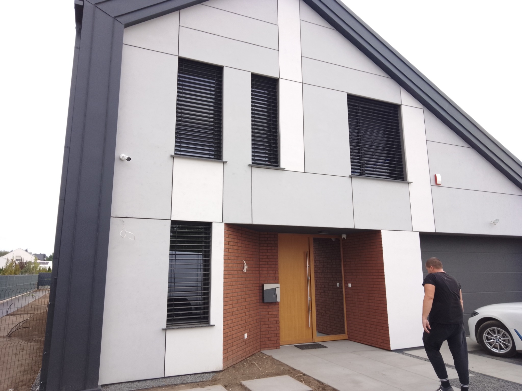 Nowoczesny dom jednorodzinny z elewacją z paneli w odcieniach szarości i białym, z ciemnymi żaluzjami zewnętrznymi i fragmentem elewacji z cegły klinkierowej, widoczna brama garażowa i osoba w tle.