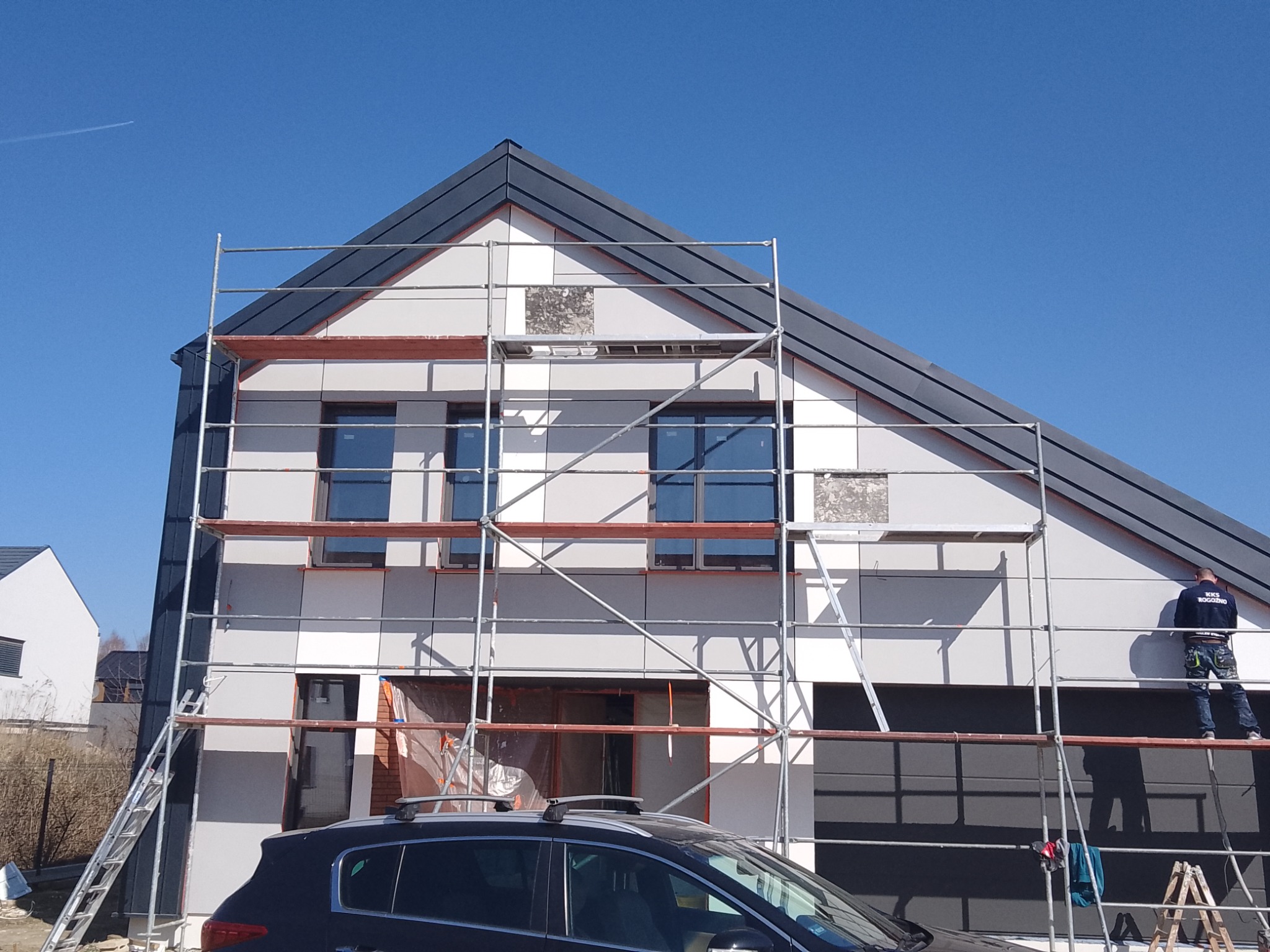 Montaż elewacji na nowym domu z użyciem rusztowania, widoczny pracownik w ciemnej bluzie i fragment zaparkowanego samochodu.