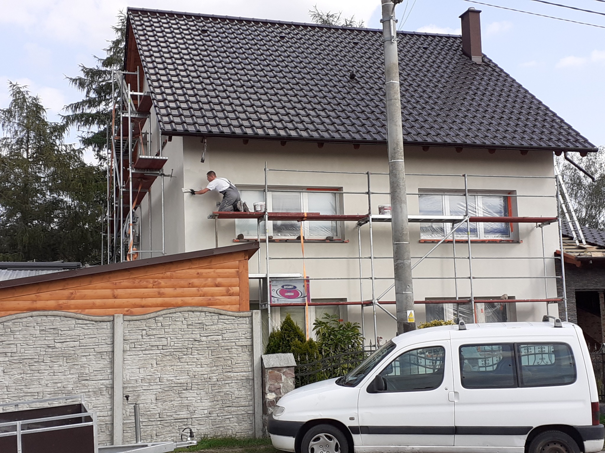 Pracownik na rusztowaniu aplikuje jasny tynk na elewacji domu jednorodzinnego, widoczny samochód dostawczy i częściowo drewniany płot.
