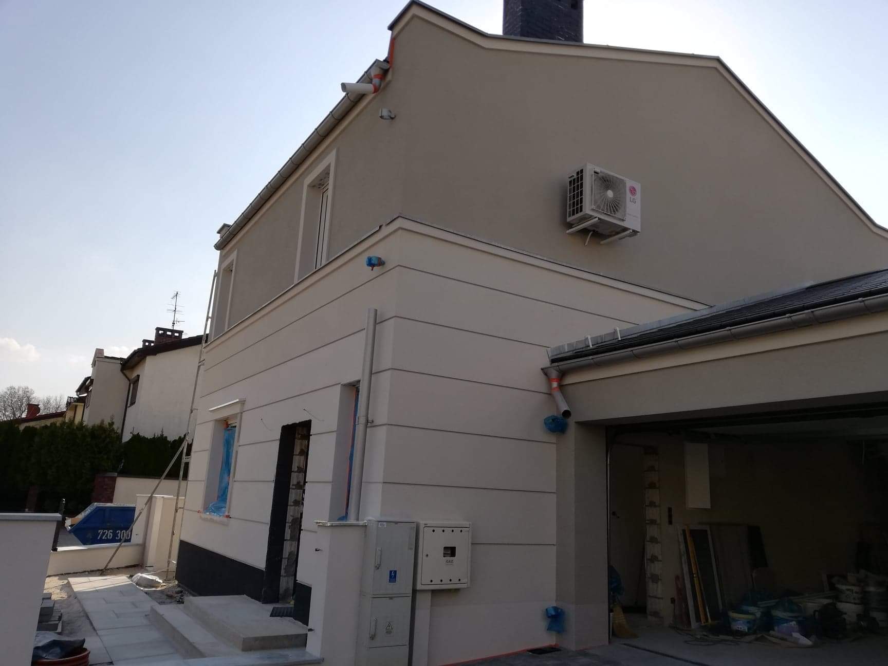 Jasna elewacja domu jednorodzinnego z widoczną jednostką zewnętrzną klimatyzacji LG, rynnami zabezpieczonymi niebieską folią i elementami wykończeniowymi wokół okien w trakcie prac.