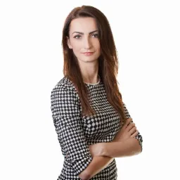 Portret kobiety z brązowymi włosami w sukience w czarno-białą kratę, z rękami skrzyżowanymi na piersiach, na białym tle.