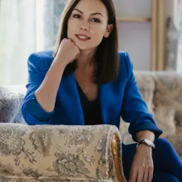 Portret uśmiechniętej brunetki w niebieskim żakiecie, siedzącej na fotelu w stylu vintage, z zegarkiem na nadgarstku i dłonią opartą o policzek.